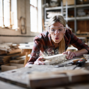 woman blowing away sawdust while wearing safety goggles